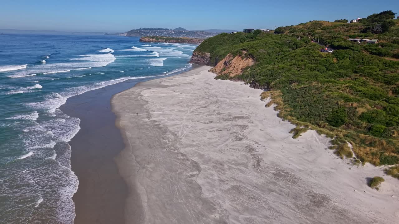 Drone flies forward above the white sand of Smaills Beach, showing ocean waves breaking along the shoreline and the green cliffs