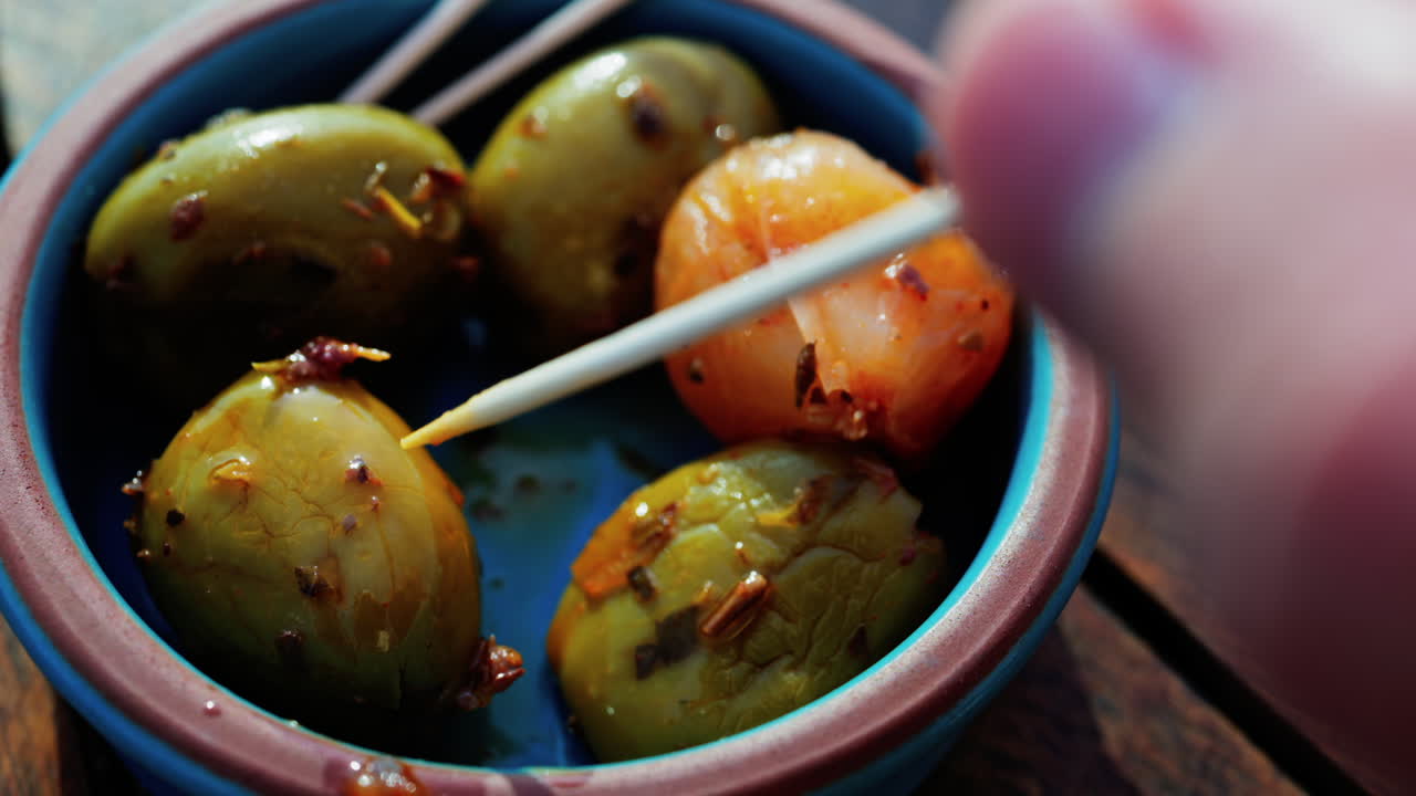 Close up of a hand picking up a green olive with a toothpick on a small bowl on a table