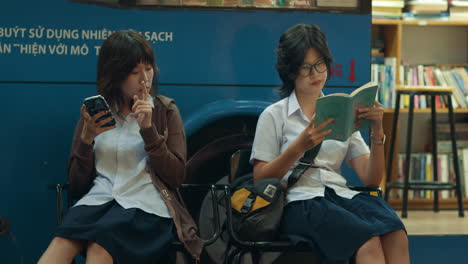 dos amigos se sientan en sillas de metal uno toca el teléfono mientras el otro lee un libro