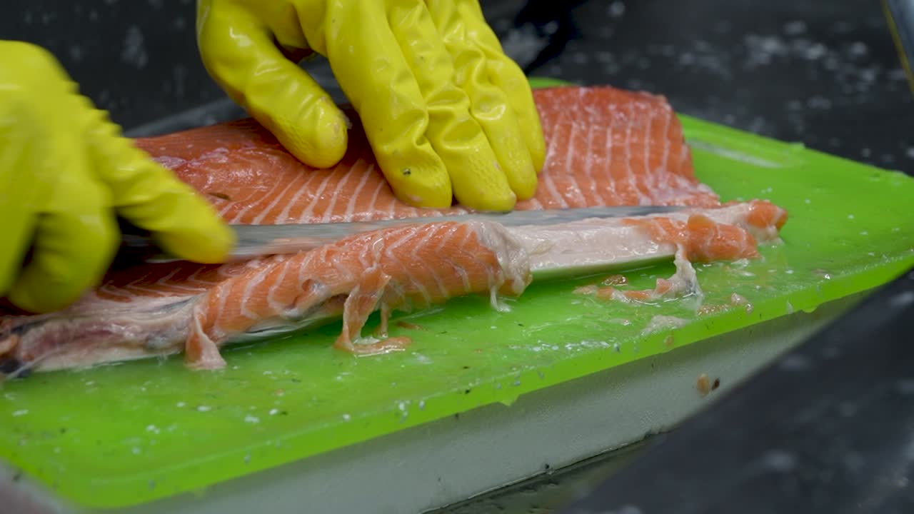 A skilled seafood preparer expertly fillets a salmon on a vibrant green cutting board. The bright yellow gloves add a pop of color as precision and care are taken in the early hours