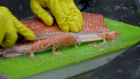 a skilled seafood preparer expertly fillets a salmon on a vibrant green cutting board. the bright yellow gloves add a pop of color as precision and care are taken in the early hours