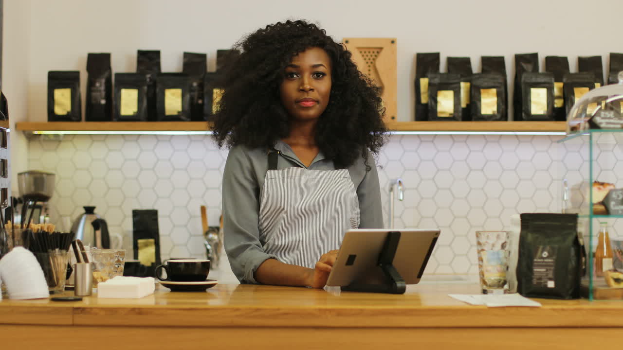 barista afroamericana con cabello rizado usando la tableta, luego mira la cámara y sonríe