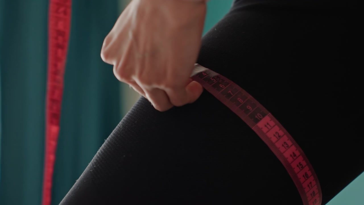Valutazione della riabilitazione della coscia a casa usando del nastro rosa, donna in leggings neri esegue un auto-controllo di tipo clinico in camera da letto, misurazione mirata per il recupero e il monitoraggio della mobilità, mani ferme, smartwatch.