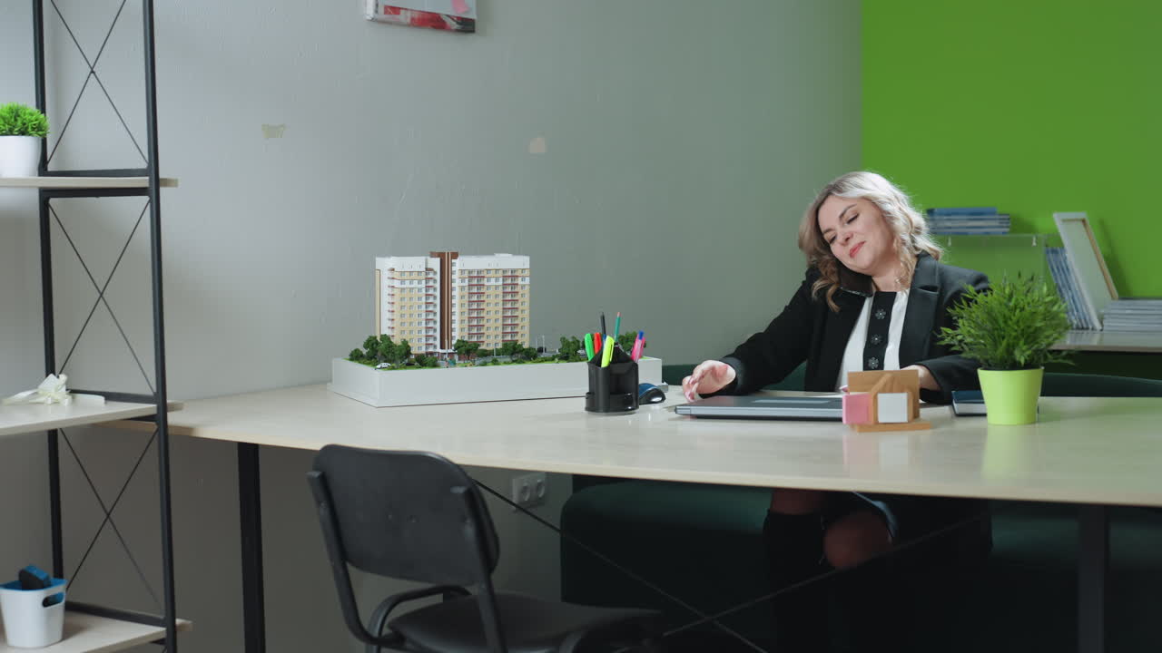 CEO on call with client opens laptop to start work in modern office with architectural model on desk, green accent wall, file shelves and organized workspace in background