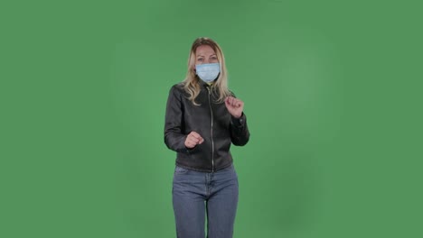 의료 마스크를 입은 아름다운 젊은 여성의 초상화는 직선적으로 보이고 재미있게 춤추고 있습니다. 검은색 재과 청바지를 입은 느슨한 머리카락을 가진 금발은 스튜디오의 녹색 화면에 있습니다. 건강 보호 코로나 바이러스 개념
