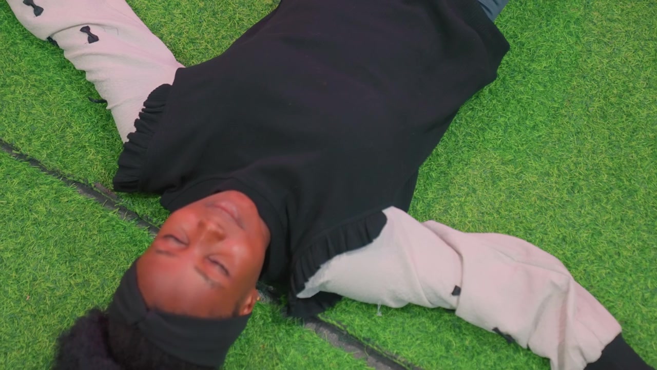 Remaja berkulit hitam melamun di rumput sintetis, potret dekat dari atas wajah santai dan lengan terbuka, sweater nyaman dan ikat kepala, latar belakang hijau pekat, ketenangan introspektif, momen gaya hidup yang alami