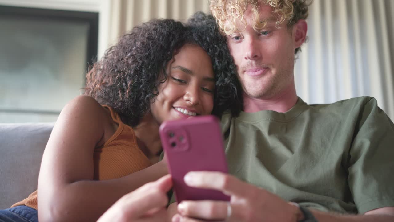 Coppia eterogenea seduta sul divano che guarda contenuti su uno smartphone con custodia magenta, scorrendo e legando.