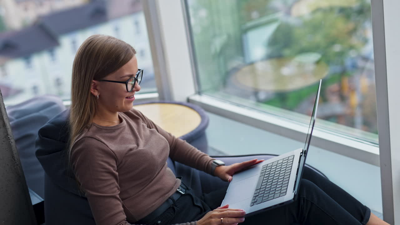 Woman in glasses opens laptop. Female starts typing on her computer sitting in comfortable bean bag chair. Side view. Blurred windows at backdrop.