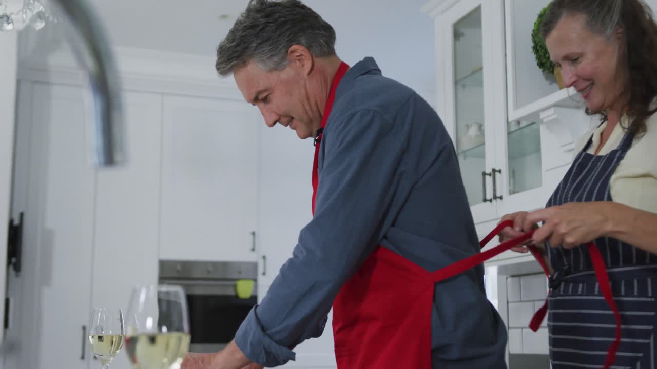 Happy senior caucasian couple in kitchen, wife tying husband's apron strings and smiling