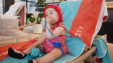 one-year old toddler on a poolside lounge chair holding tumbler of water and playing with blue strap in her mouth