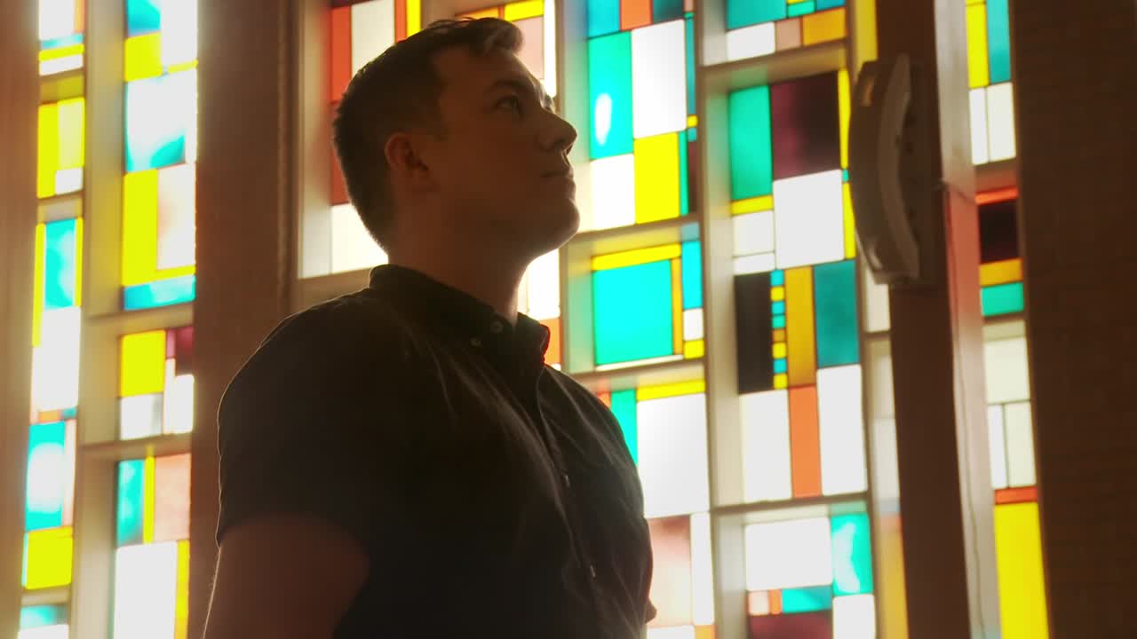 Close up of a young ethnic male inside a church sanctuary who is calmly mediating in an isolated and faith-building posture in front of a gorgeous stained glass window wall aglow in the sun