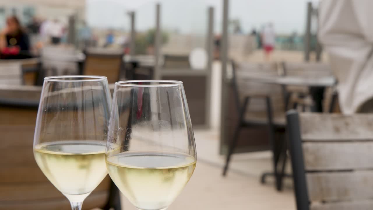 Two white wine glasses on seaside cafe table as waiter approaches, natural daylight, shallow focus