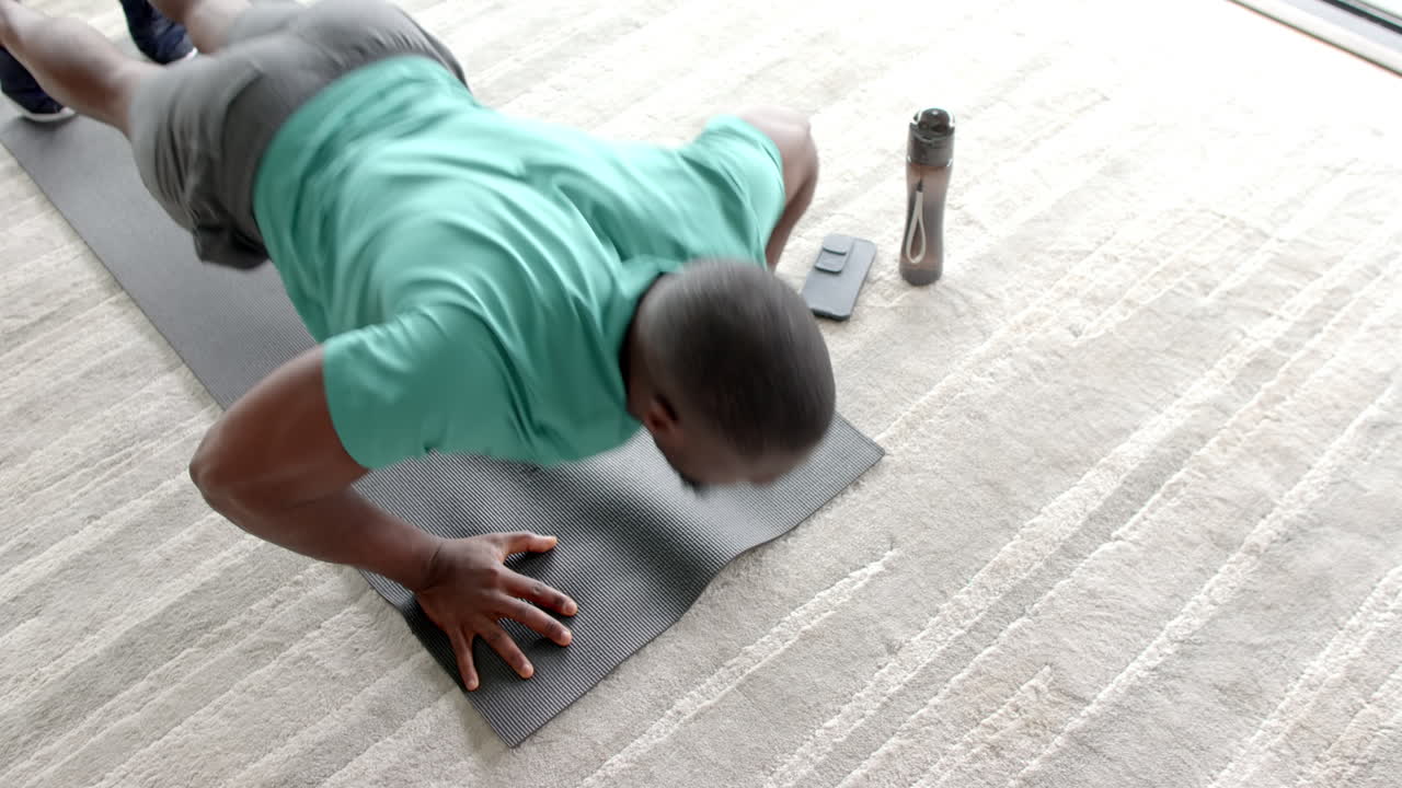 Doing push-ups on yoga mat, African American man exercising at home with water bottle