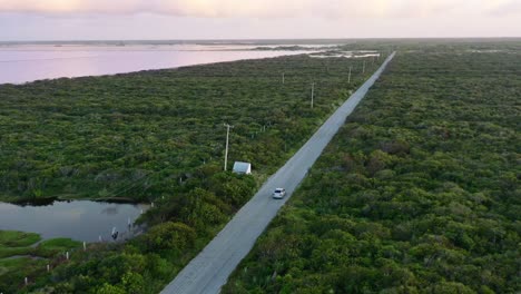 Coche-Conduciendo-Hacia-La-Puesta-De-Sol-En-Una-Larga-Carretera-Vacía-Rodeada-De-Exuberante-Vegetación-Y-Lago-En-Las-Coloradas-México,-Antena