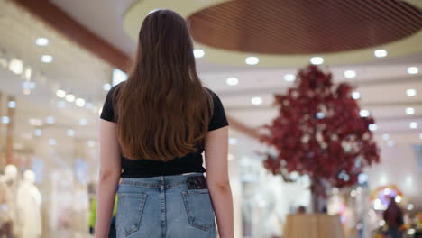 검은 과 청바지를 입은 여성이 현대 쇼핑몰을 통해 자신감 있게 고, 뒷 주머니에 보이는 전화, 따뜻한 조명과 배경에 흐릿한 구매자를 가진 세련된 소매 공간