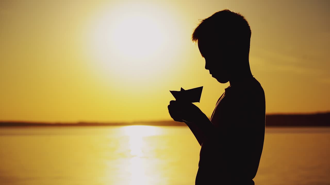 Young boy is making a paper ship standing near the river at the evening sunset. Silhouette of the boy playing with paper boat outside