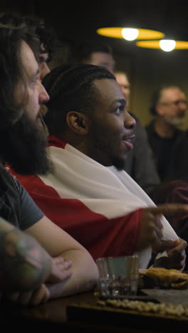 vertical shot of african american fan celebrating with friends when football team scores goal and wins the tournament. group of multicultural friends watch live soccer match sitting in sports bar or pub.