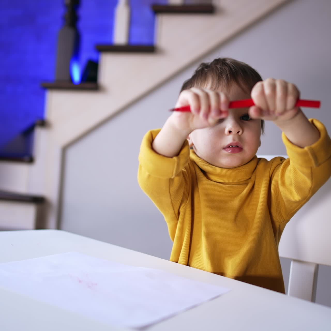 Small toddler in yellow sweater sitting at desk. Kid holds a red pen and waving it. Child drawing at home