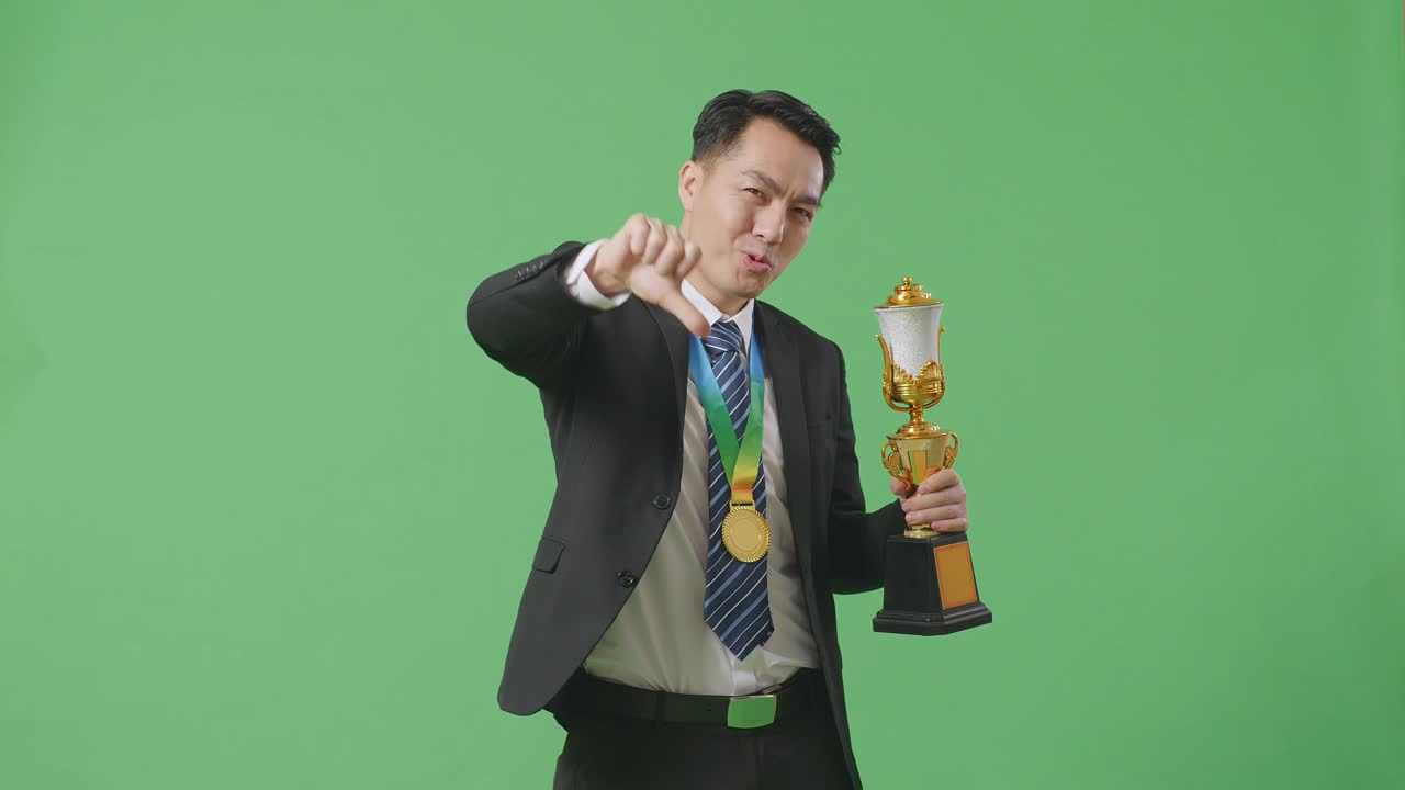 슈트와 타이를 입은 아시아 사업가, 금메달과 트로피를 들고, 엄지손가락을 아래로 가리키며, 스튜디오의 녹색 스크린 배경에서 패배자를 조롱하는 몸짓