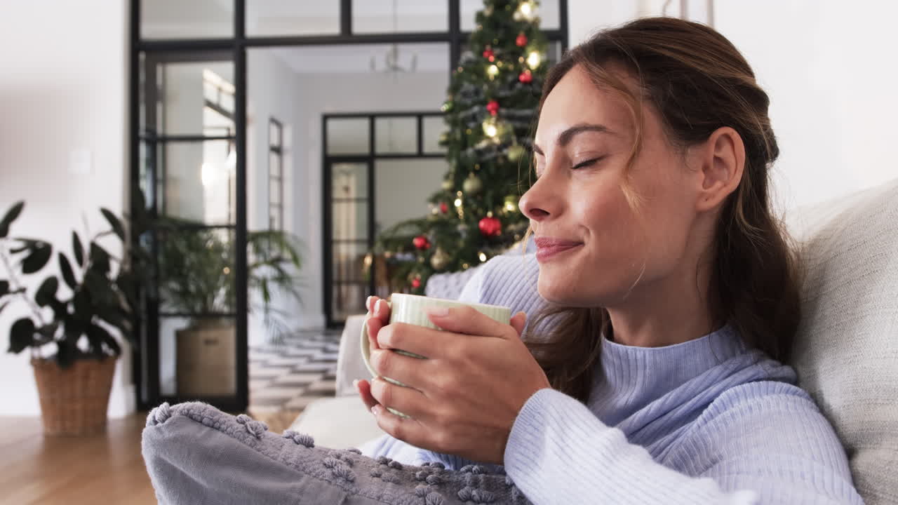 Relaxing at home, woman enjoying hot drink by Christmas tree, feeling cozy