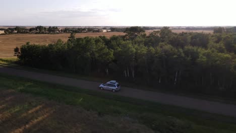 Sideways-following-aerial-shot-of-silver-minivan-driving-along-dusty-gravel-road-in-rural-Alberta