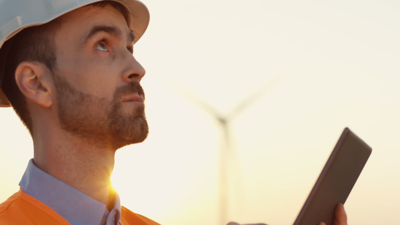 Close-up view of caucasian male engineer in helmet and uniform using tablet at wind station of renewable energy