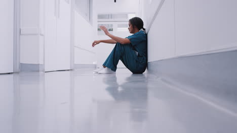 stressed and overworked female doctor wearing scrubs sitting on floor in hospital corridor