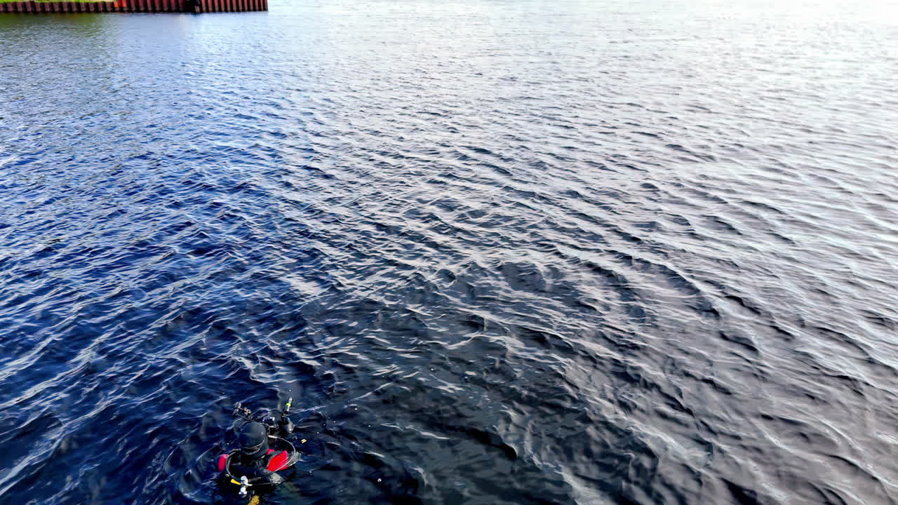 An oblique drone tracks a lone diver towing a bright safety float across rippled harbor water, emphasizing inspection training, awareness, and steady endurance against the textured surface