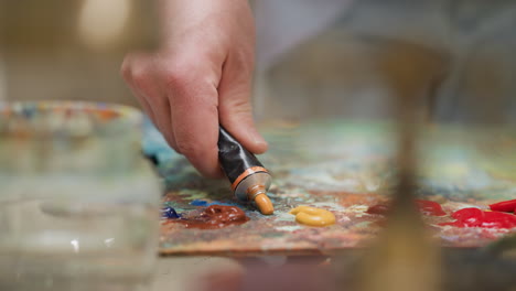 creator squashes paint out of metal tube at desk closeup. skilled artist squeezes out orange acrylic pigment for artwork in shop. painter supplies