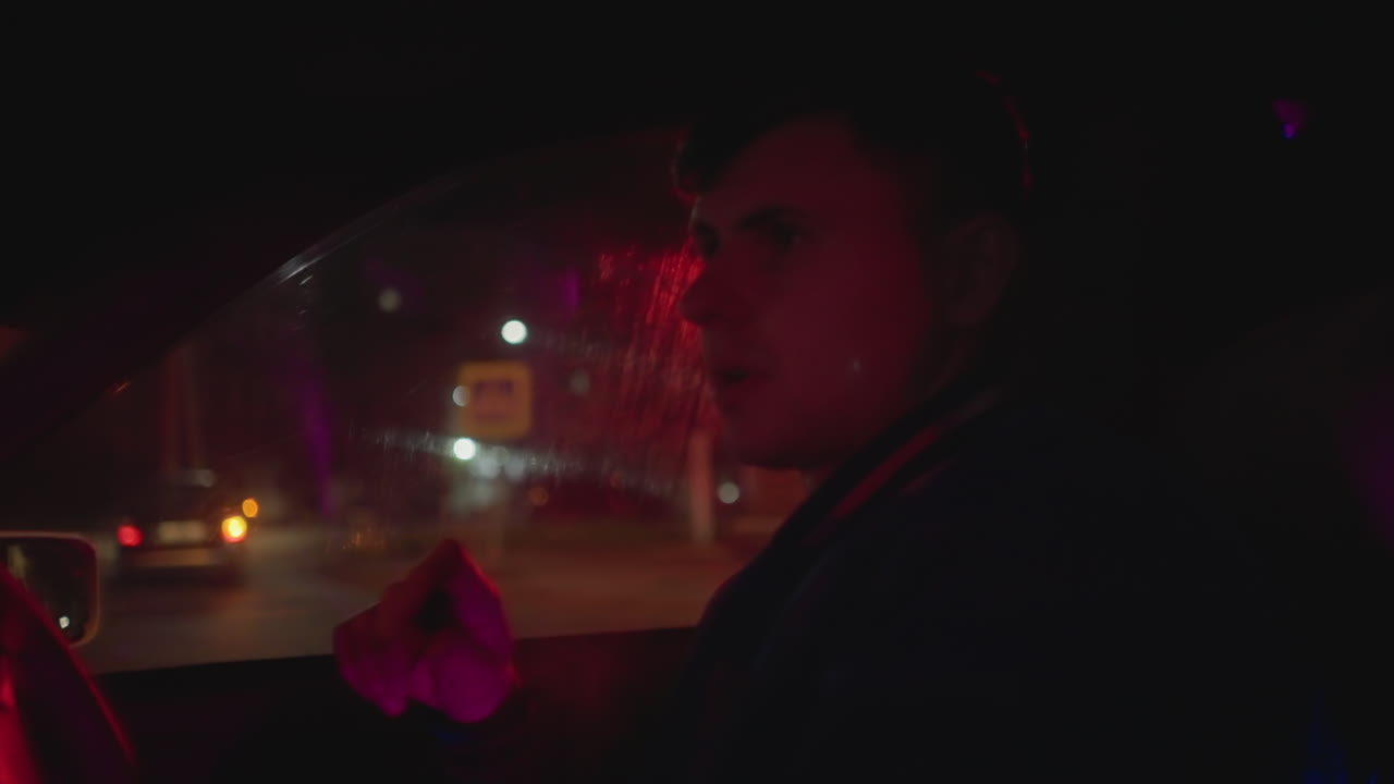 밤에 전자담배를 피우는 남성 운전자, 네온 불빛이 비치는 차 안에서 생각에 잠긴 차량 공유 운전자가 빨간불에 멈춰 서서 손에 든 전자담배가 보이고, 대시보드 불빛과 도시 보케가 창문을 통해 보이며, 그림자가 드리워진 옆모습