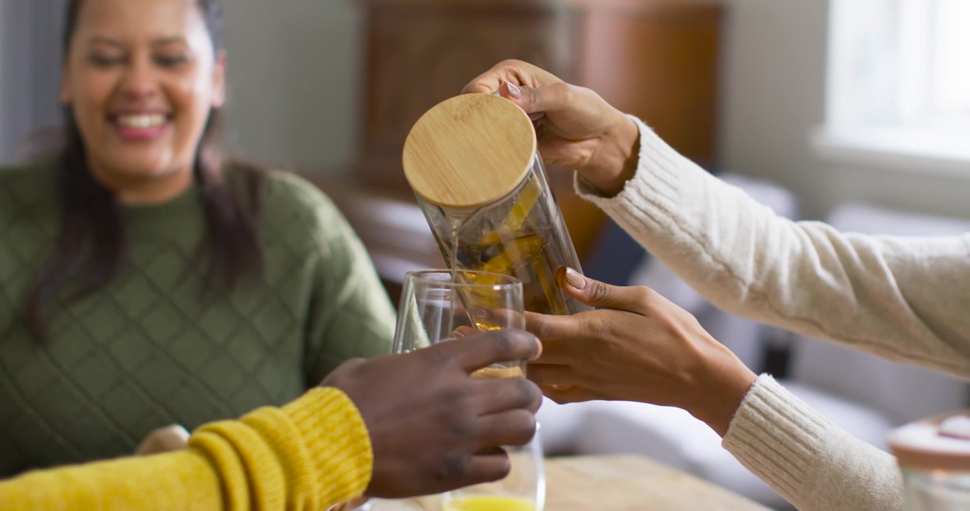 Mulher afro-americana adulta sorrindo à mesa enquanto uma pessoa serve uma jarra, preenchendo o copo vazio no início da refeição.