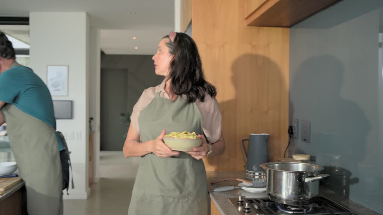Coppia con grembiuli che cucina in cucina usando una ricetta su tablet, mentre tagliano, si passano una ciotola e mescolano una pentola.
