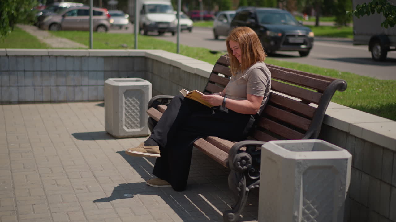 Side view of pupil sitting on wooden bench reading and flipping page of book under bright sunlight, calm posture and focused look showing learning mood, soft blur background