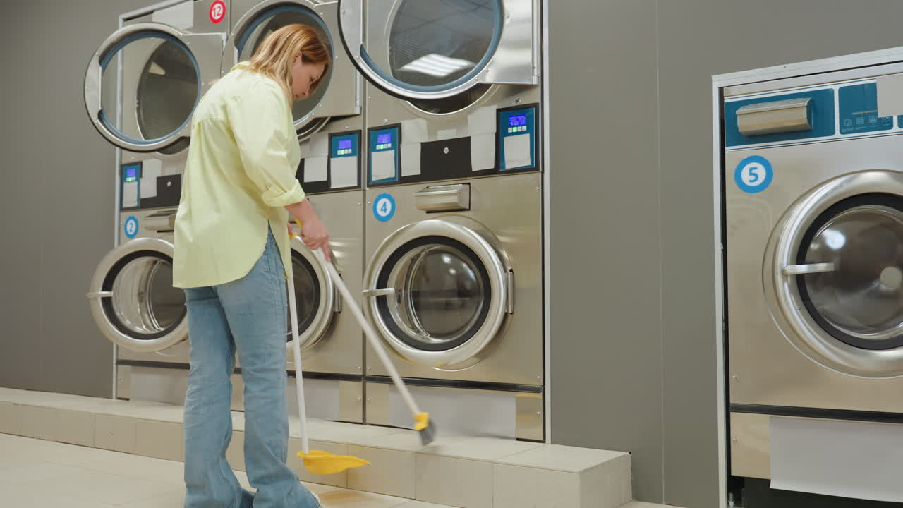 Back view staff in yellow shirt walks toward industrial washers, sweeps ground with broom and dustpan, maintaining bright laundromat with stainless machines and tiled floor, routine cleaning scene