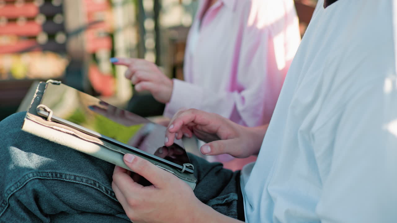 primer plano de una mujer con una camiseta rosa y uñas pulidas operando una tableta mientras continúa escribiendo en su computadora portátil, sentada al aire libre con un hombre escriviendo en su propio dispositivo