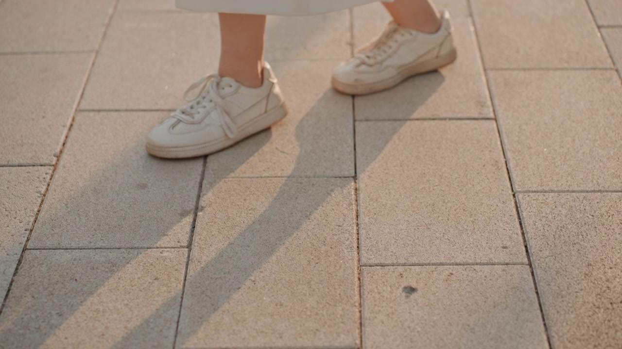 Las piernas de una mujer y zapatillas blancas se mueven dinámicamente sobre un pavimento de piedra iluminado por el sol, capturando el movimiento de caminar o bailar. El enfoque permanece en su parte inferior del cuerpo con una iluminación suave que realza la textura.