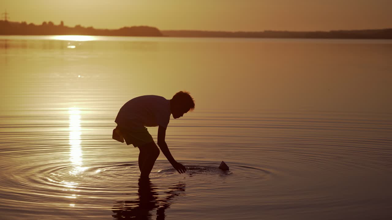 Boy near the lake at sunset. Silhouette of a boy in water with paper boat. Child putting origami ship on the beautiful evening river background. Freedom concept.