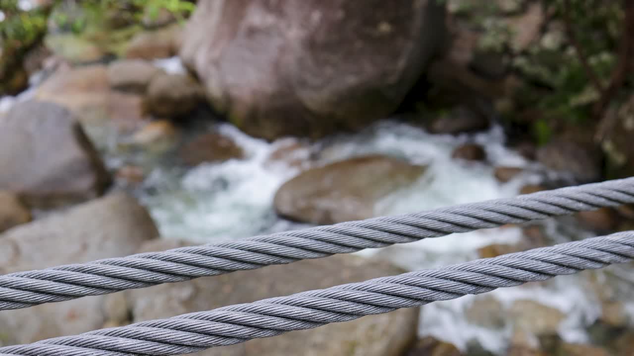 A sturdy steel cable stretches across a rocky river in a forest setting, with water flowing rapidly below