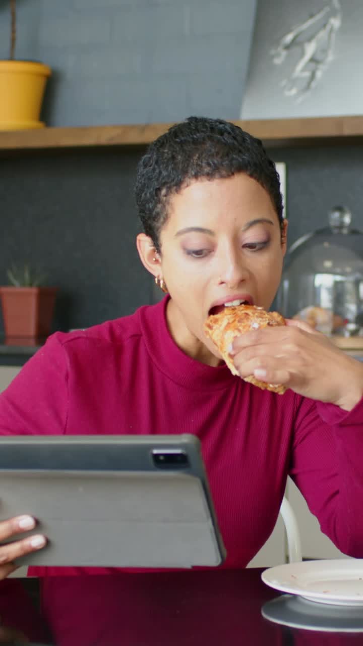 Vertical video: Reading tablet, woman picking and eating pastry at kitchen table in burgundy top