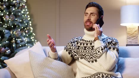 close up portrait of joyful young man wearing xmas sweater sitting in room near decorated glowing christmas tree speaking on cellphone telling holiday greetings