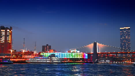 riverboats move by the east river at night. pier 17 bar and the brooklyn bridge at backdrop. new york, usa