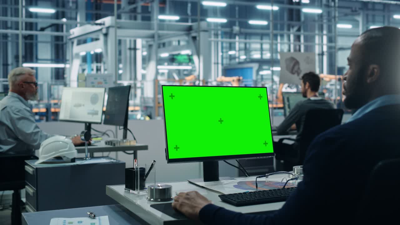 자동차 공장 사무실: 흑인 남성 자동차 수석 엔지니어가 그의 책상에 앉아 녹색 화면 크로마 키 컴퓨터에서 일하고 있습니다. 자동 로 팔 조립 라인 제조. 어깨에서 중간.