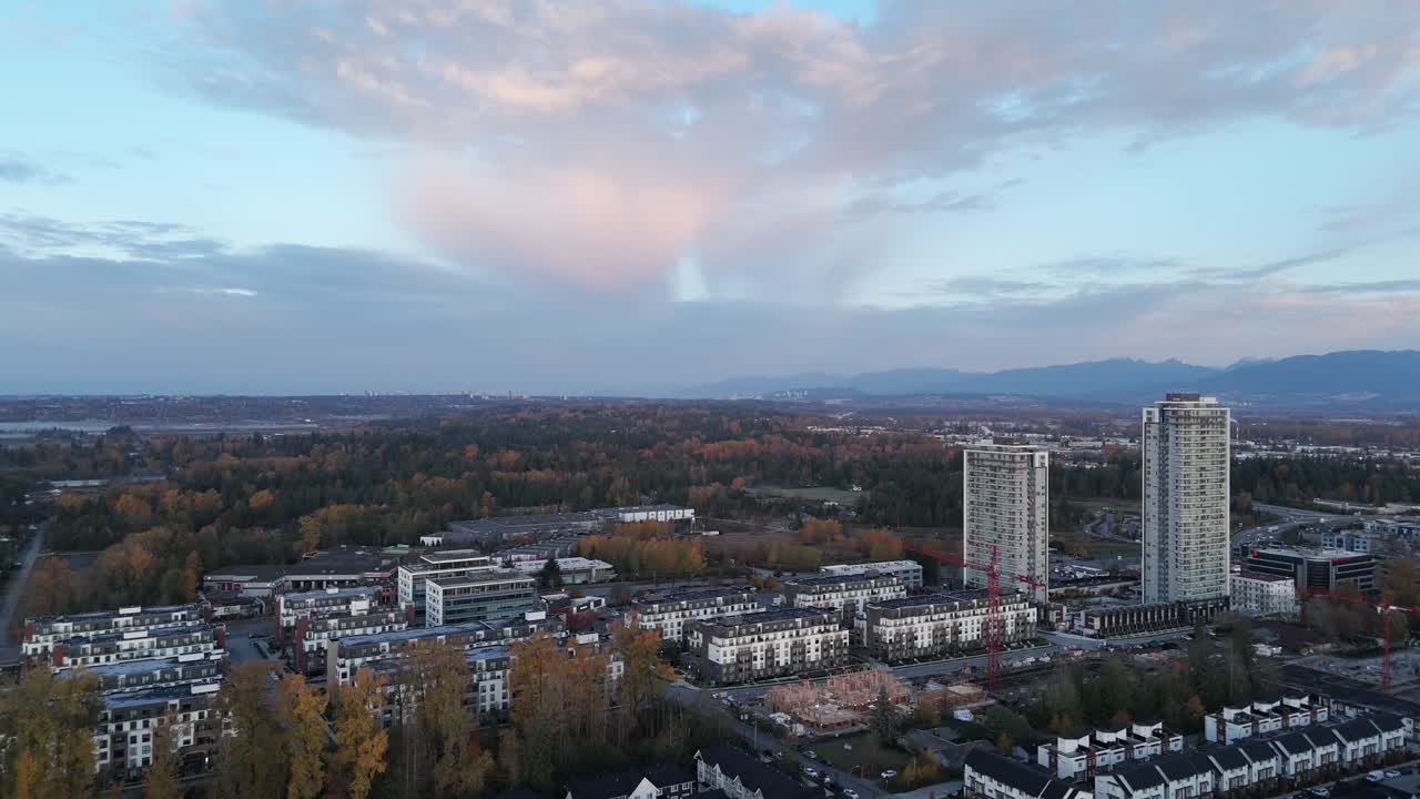 Carvolth, Langley City, Kanada – Moderneja pilvenpiirtäjiä ja asuinalueita syksynväristen puiden keskellä, kaukaiset vuoret pehmeän pastellitaivaan alla – Drone lentää eteenpäin
