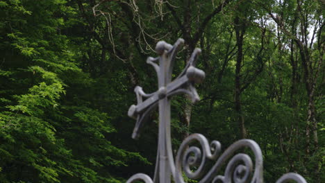 Cast-iron-cross-on-ornate-gate-top,-trees-of-deep-forest-behind