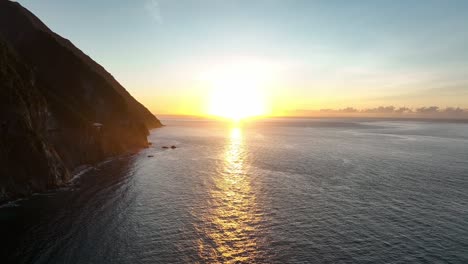 Aerial-establishing-shot-of-golden-sunset-shining-on-ocean-water-beside-mountain-coastline-in-Asia---Panorama-view