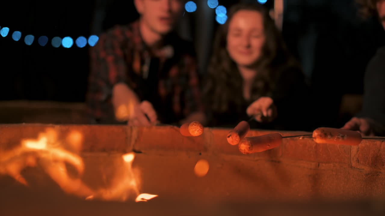 A group of happy young friends near a campfire at glamping, night. Frying sausages. Slow motion