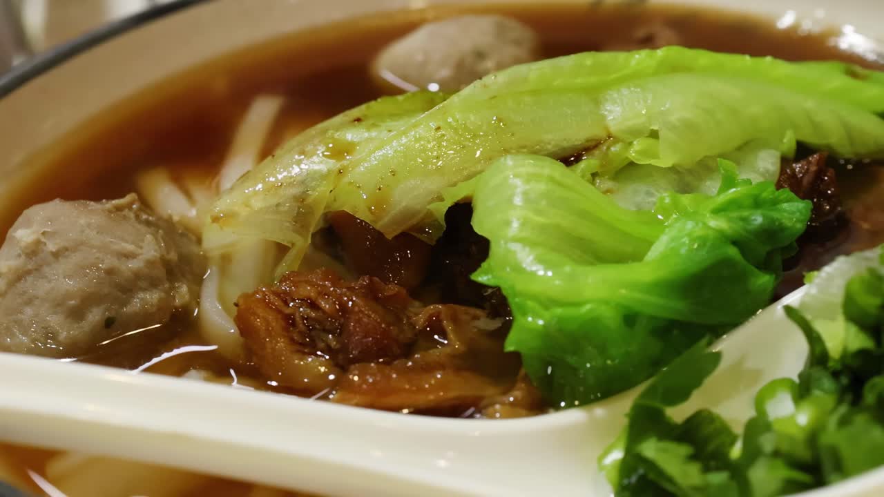 A detailed view of beef noodle soup featuring meatballs, lettuce, and a rich broth in a spoon.