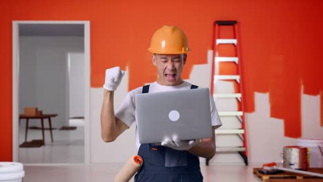 pintor hombre asiático con casco de seguridad mirando la computadora portátil luego gritando meta celebrando mientras estaba de pie con la pared de pintura en la habitación