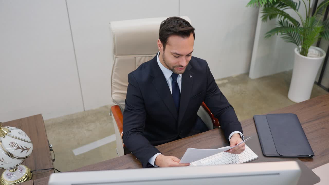 A businessman sits at a desk in an office reviewing paperwork documents or reports - high angle view
