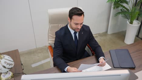 a businessman sits at a desk in an office reviewing paperwork documents or reports - high angle view